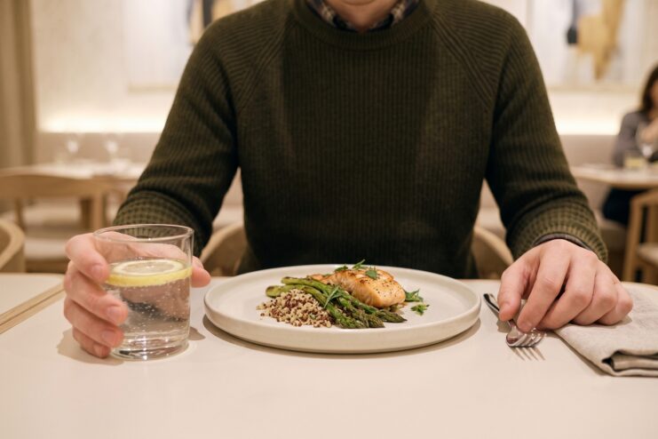 Primer plano de las manos de un hombre en la mesa junto a un plato de pescado y un vaso de agua con una rodaja de limón.