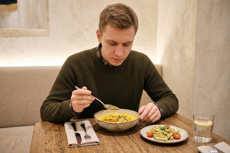 Hombre tomando sopa con una cuchara en una cafetería, con un plato de macarrones al lado sobre la mesa.