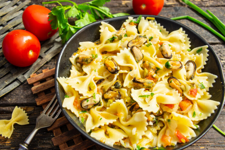 Plato hondo oscuro con farfalle y mejillones en salsa de tomate sobre una mesa de madera.