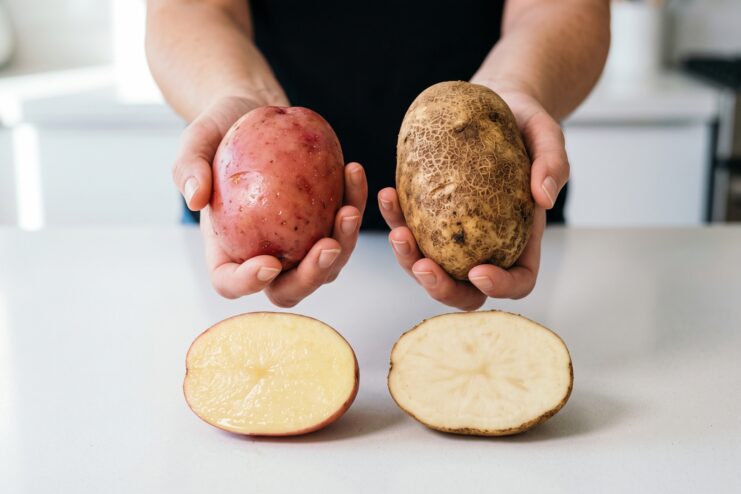 Comparativa de una patata roja cerosa y otra marrón harinosa, ambas cortadas por la mitad sobre una mesa blanca.