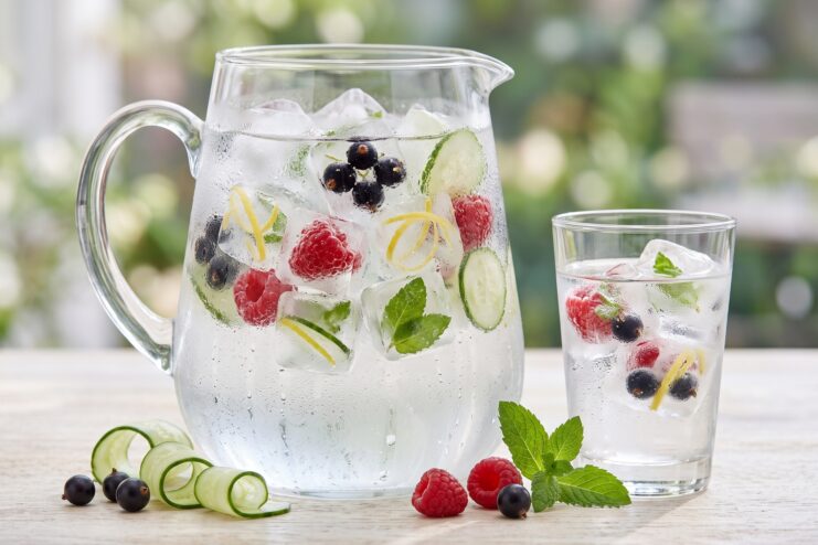 Agua Refrescante Con Vistoso Hielo De Frutas Y Verduras Jarra y vaso de cristal con agua y hielo que contiene frutos rojos, rodajas de pepino y hojas de menta.